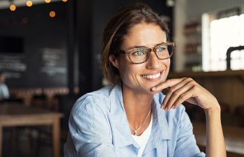 Woman wearing glasses smiles, sitting in a cafe with a relaxed posture.